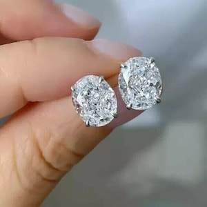 Puede incluir: Un par de pendientes de diamantes con corte ovalado. Los diamantes están engastados en un metal de color plateado y brillan intensamente. Los pendientes se sostienen sobre un fondo borroso.