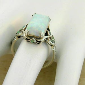 May include: A silver ring featuring a large, rectangular opal gemstone. The opal has a milky white appearance. The ring is accented with small green gemstones and intricate silver detailing on the band.