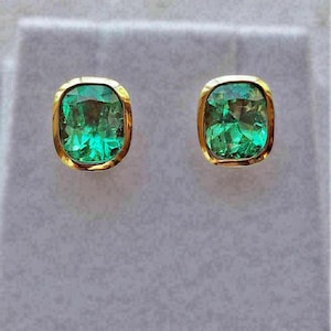 May include: A pair of square-cut emerald earrings with a gold-coloured bezel setting. The emeralds are a vibrant green colour, set against a grey background. The earrings are a classic jewellery item.