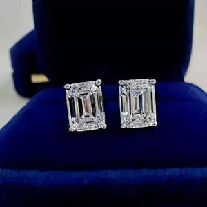 Puede incluir: Un par de brillantes pendientes de diamantes con corte esmeralda. Las piedras claras y facetadas están engastadas en una montura de plata u oro blanco, presentadas sobre una caja de joyería de terciopelo azul oscuro. Una joya clásica.