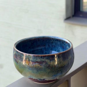 Op de afbeelding: Een ronde keramische kom met een reactief glazuur in gemengde kleuren van diepblauw, bosgroen en aards bruin. De binnenzijde heeft een glanzende donkerblauwe afwerking, terwijl de buitenzijde verloopt in gelaagde tinten groen en bruin.