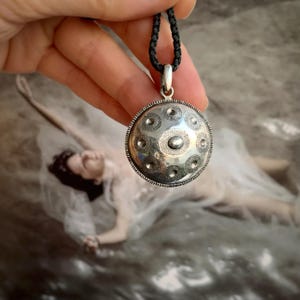 Könnte beinhalten: Eine silberne Anhänger Halskette mit einem kreisförmigen, gemusterten Design. Der Anhänger hat eine Reihe von kreisförmigen und Punktmustern mit einer zentralen ovalen Form. Die Halskette hat eine schwarze Kordel und einen silbernen Verschluss.