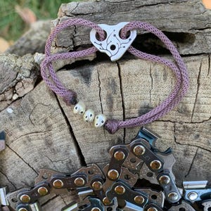 Könnte beinhalten: Ein lilafarbenes Kordelarmband mit einem silbernen Anhänger in Form eines Herzens mit einem Blumendesign. Das Armband hat vier silberne Perlen.