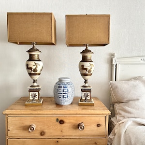 May include: Two table lamps with rectangular tan fabric shades and urn-shaped bases featuring gold and white classical chariot motifs on square pedestals, positioned on either side of a round blue and white porcelain ginger jar with a lid.