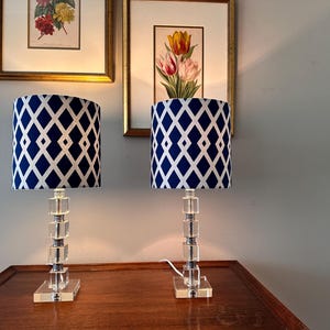 May include: Two matching table lamps with clear, stacked crystal bases and navy blue and white diamond patterned shades. The lamps are on a dark wood surface, with framed floral art in the background.
