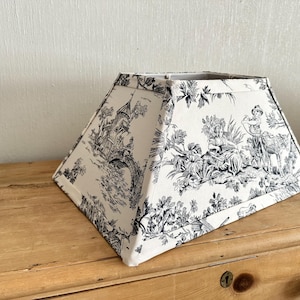 May include: A rectangular lamp shade featuring a black and white toile de jouy pattern. The fabric displays detailed illustrations of pastoral scenes, including figures, trees, and architectural structures, finished with matching fabric trim.