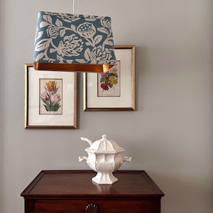May include: A teal and white floral lampshade with a copper-colored trim hangs above framed botanical prints and a white ceramic soup tureen on a dark wood table. The lampshade is the focal point, adding a touch of elegance to the room.