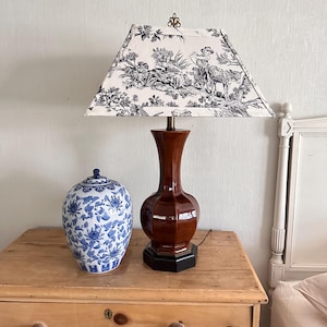 May include: A decorative table lamp with a brown, glossy base and a black base. The lampshade is white with a black toile pattern. A blue and white floral ceramic jar sits next to the lamp on a wooden surface.