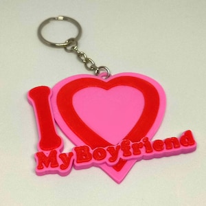 May include: A pink and red heart-shaped keychain with the words "I My Boyfriend" in red. The keychain has a silver metal ring and chain. The design is a romantic gift.