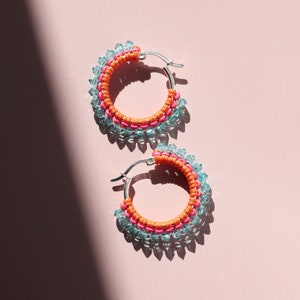 May include: A pair of hoop earrings with a silver metal hoop and a beaded design. The beads are a mix of light blue, pink, and orange.