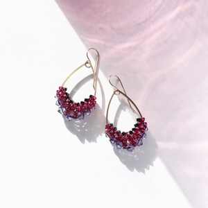 May include: A pair of teardrop-shaped earrings with a gradient of purple and pink crystals. The earrings are made with gold-toned wire and have a dangling design.