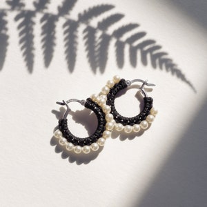 May include: A pair of hoop earrings with a black woven design and white pearl accents. The earrings are set against a white background with the shadow of a fern leaf. The earrings have a silver clasp.