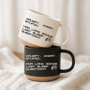 May include: A stack of two ceramic mugs. The top mug is off-white with black text, and the bottom mug is matte black with white text. Both feature the printed words: CRUMPY. ANGRY. STUPID. HOW LONG SINCE LAST SLEEP. QUESTION? and a small robot icon.