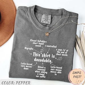 Op de afbeelding: Een peperkleurig t-shirt met witte tekst: This shirt is decodable, omringd door taalkundige notities waaronder Closed Syllable, Digraphs, r-controlled, s says /z/, Latin-based en Silent-e. Een dunne gouden ketting rust op de kraag.