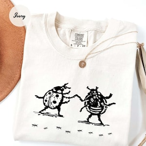 May include: Ivory t-shirt with a black illustration of two beetles holding hands, with ants below. The shirt has a crew neck and a "Comfort Colors" tag. A gold necklace with a pendant is draped on the shirt.