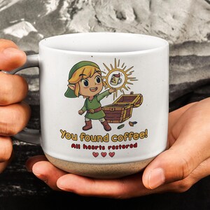 May include: A white ceramic mug with a speckled base featuring a cartoon character in a green tunic holding a glowing coffee mug above an open treasure chest. The text reads "You found coffee! All hearts restored" with three red heart icons below.