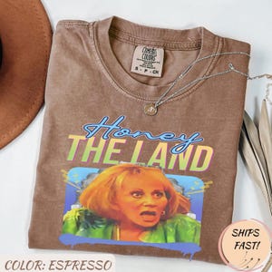 May include: Espresso-coloured Comfort Colors t-shirt with a graphic of a woman and the text "Honey The Land". The shirt has a crew neck and short sleeves. A silver necklace with a pendant is draped on the shirt.