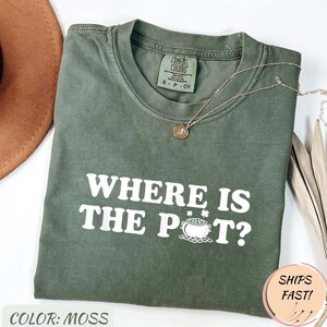May include: A moss green t-shirt with the text "WHERE IS THE POT?" in white. The shirt has a crew neck and short sleeves. A gold necklace is draped across the shirt. The shirt is labeled "Comfort Colors".