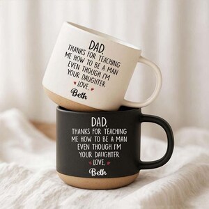 May include: Two ceramic mugs stacked vertically, one cream-colored and one black. Both feature the text: DAD, THANKS FOR TEACHING ME HOW TO BE A MAN EVEN THOUGH I'M YOUR DAUGHTER LOVE, Beth. Each mug has a natural, unglazed tan base and a side handle.