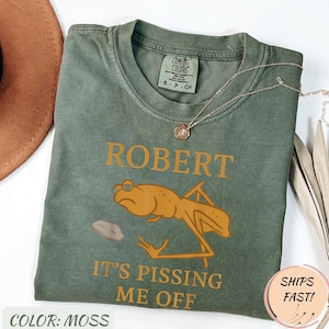 Könnte beinhalten: Moosgrünes T-Shirt mit dem Text "ROBERT" über einer Cartoon-Froschgrafik und dem Satz "IT'S PISSING ME OFF". Das Shirt hat einen Rundhalsausschnitt und kurze Ärmel. Eine silberne Halskette mit Anhänger liegt über dem Shirt.