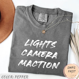 May include: Pepper-colored t-shirt with the words "LIGHTS CAMERA ACTION" in white script. The shirt has a crew neck and short sleeves. A silver necklace with a small pendant is draped on the shirt. The shirt is folded on a white surface.