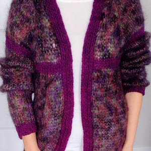 May include: A long, open-front cardigan in shades of purple, pink, and green. The sweater features a deep purple trim along the edges, cuffs, and pockets. The sleeves are long and the knit has a textured, fuzzy appearance.