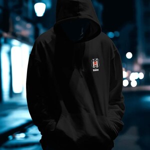 May include: Black hooded sweatshirt with a front pocket and a small emblem on the chest. The emblem features a crest design with a red and white color scheme and the word "Name" below it.