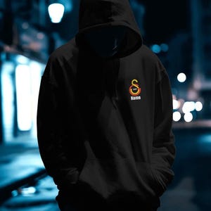 May include: Black hooded sweatshirt with a front pocket and drawstring. The sweatshirt features a logo with the text "Name" and the year "1905" in a circular design. The background is a blurred night scene.