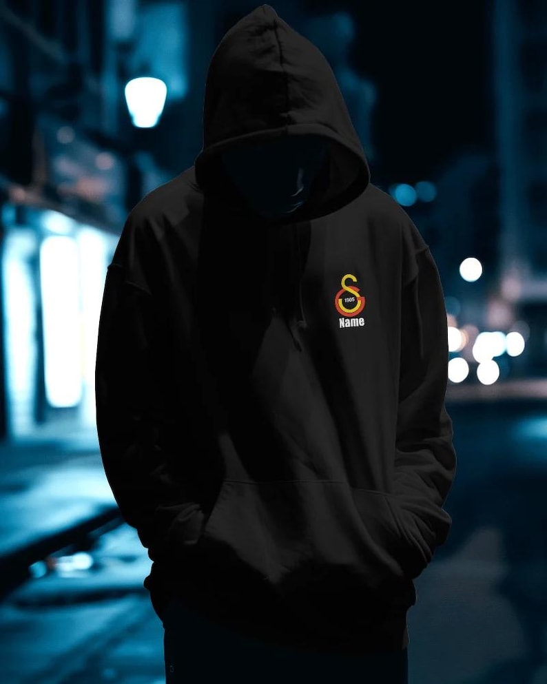 May include: Black hooded sweatshirt with a front pocket and drawstring. The sweatshirt features a logo with the text "Name" and the year "1905" in a circular design. The background is a blurred night scene.