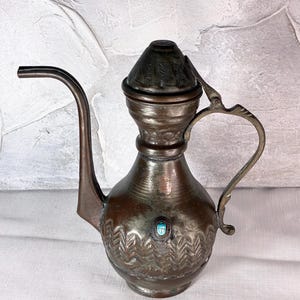 May include: A copper-toned metal ewer featuring a long, curved spout, a tapered neck, and a hinged lid. The body is decorated with embossed zigzag patterns and a single oval-shaped turquoise-colored inlay centered on the front surface.