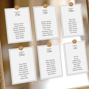 May include: A framed seating chart with six cards, each labeled with a table number. The cards are white with gold accents and feature guest names. The frame is gold, and the cards are held in place with gold wax seals.