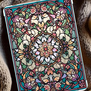 Può includere: Una custodia per tablet con un design a vetrata raffigurante motivi floreali e geometrici nei toni del rosa, verde, giallo e bianco. La custodia ha un bordo turchese e una cornice color oro. La custodia è appoggiata su una coperta di maglia beige.