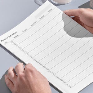 May include: A white weekly schedule notepad with a time grid and days of the week printed in black. The notepad is held by hands and is on a light gray surface. The words "Weekly Schedule" are printed at the top.