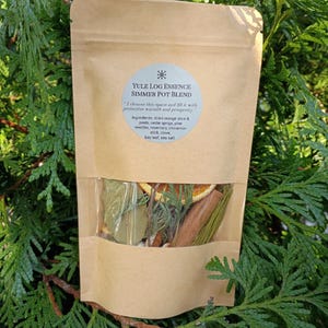 May include: A brown paper pouch containing a Yule Log Essence Simmer Pot Blend. The label lists ingredients including dried orange, cedar, pine needles, rosemary, cinnamon, clove, bay leaf, and sea salt. A clear window reveals the dried contents.