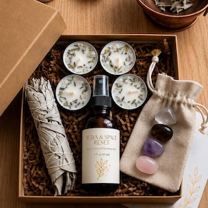 May include: A ritual kit in a cardboard box containing four small round candles with botanical inclusions, a 2 fl oz amber glass spray bottle labeled Aura & Space Reset, a bundle of dried sage, and a beige drawstring pouch with four polished stones.