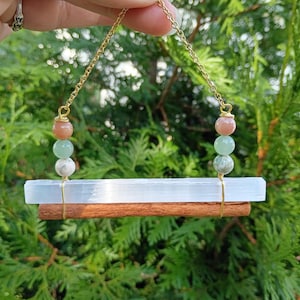 May include: A hanging ornament featuring a horizontal white selenite crystal wand paired with a brown cinnamon stick. The two pieces are connected by gold-toned wire and chains, accented with spherical peach, light green, and white marbled beads.