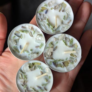 May include: Four small, round, white candles with silver rims. Each candle is decorated with small pieces of clear crystals, green herbs, and lavender. The candles are held in a person's open palm.