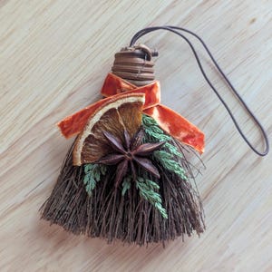 May include: A small decorative broom ornament crafted from dark brown twigs, featuring a dried orange slice, a star anise, and a sprig of green cedar. An orange velvet ribbon is tied around the handle, which includes a brown hanging cord.