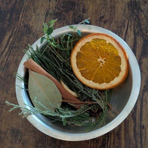 May include: A silver bowl filled with dried orange slices, cinnamon sticks, bay leaves, and sprigs of herbs. The arrangement is set against a dark wooden surface, creating a rustic and natural aesthetic.