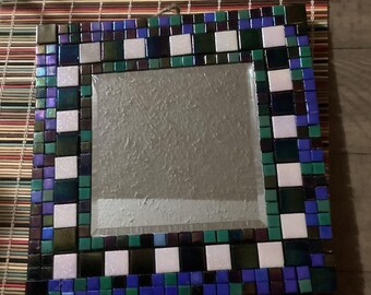 May include: Square mosaic mirror with a decorative border. The border features a pattern of white, green, blue, and purple tiles. The mirror is displayed on a striped bamboo mat.