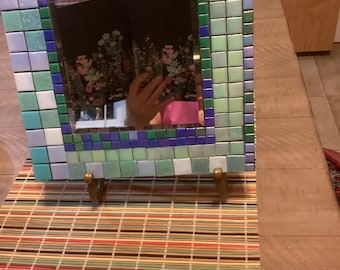 May include: Square mosaic mirror with a frame of small, colorful tiles in shades of blue, green, and white. The mirror is propped up on small, gold-colored stands. The reflection shows a floral pattern.