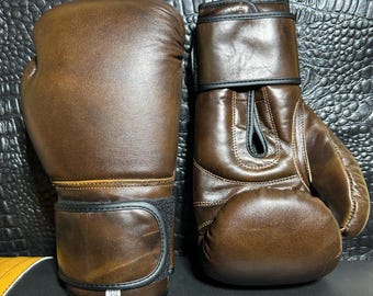 May include: A pair of brown leather boxing gloves. The gloves have black accents on the wrist straps and seams. The leather has a distressed appearance. The gloves are designed for protection and impact absorption during training or competition.