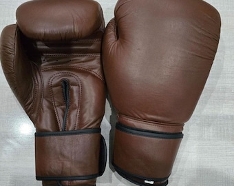 May include: A pair of brown leather boxing gloves. The gloves are a dark brown colour with black wrist straps. These gloves are designed for boxing or other combat sports, offering protection and impact absorption.
