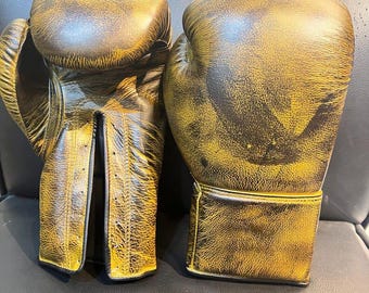 May include: A pair of boxing gloves in a distressed gold and brown color scheme. The gloves are made of leather and show signs of wear, with visible stitching and padding. The gloves are designed for protection and impact absorption during training or competition.