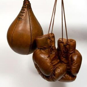 Puede incluir: Un saco de velocidad de cuero marrón y un par de guantes de boxeo de cuero marrón cuelgan de una viga de madera. El saco de velocidad tiene forma de lágrima y está cosido. Los guantes están atados y desgastados.