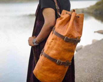 Könnte beinhalten: Ein großer, zylindrischer, orange-brauner Lederrucksack mit braunen Riemen und Schnallen. Die Tasche hat einen Kordelzugverschluss oben und wird über der Schulter getragen. Die Tasche ist zum Tragen von Gegenständen konzipiert.