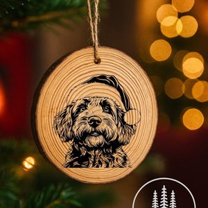 May include: A circular wooden Christmas decoration featuring a laser-etched image of a dog in a Santa hat. The ornament is made from light-coloured wood, showing a natural grain pattern. A jute twine loop is attached for hanging.