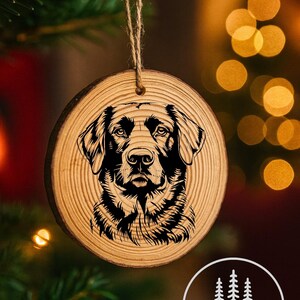 May include: A round wooden ornament with a detailed black engraving of a dog's face. The ornament hangs from a twine string. The background is blurred with Christmas tree branches and bokeh lights. The ornament has the text "WestCoast Wood Working & Laser-Art".