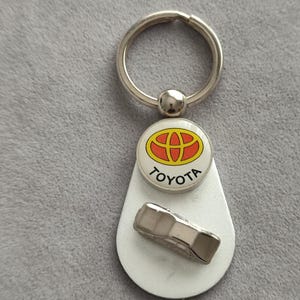 Könnte beinhalten: Ein silberfarbener Metallschlüsselanhänger mit einem runden weißen Emblem, das das rote und gelbe Toyota-Logo sowie das Wort TOYOTA in schwarzer Schrift zeigt. Unter dem Emblem ist eine kleine, dreidimensionale silberne Autofigur befestigt.