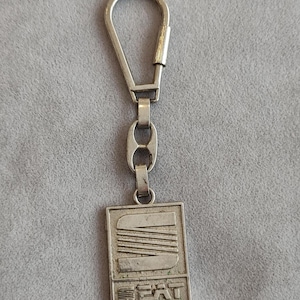 Peut inclure: Un porte-clés en métal argenté doté d'un pendentif rectangulaire avec le logo SEAT en relief sur la partie supérieure et le mot SEAT en lettres capitales sur la partie inférieure. Le pendentif est relié à un anneau en forme de goutte.